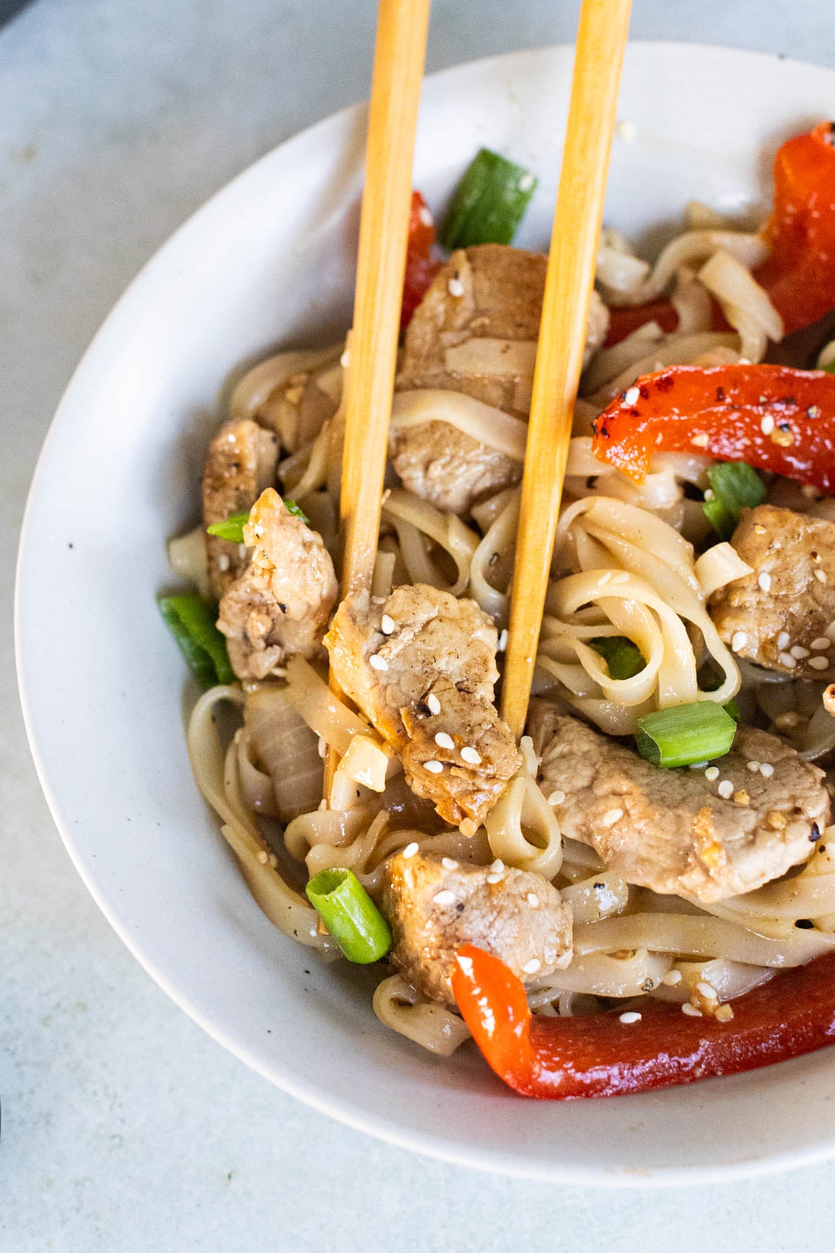 A bowl of noodle stir-fry with pieces of pork tenderloin, red bell pepper, green onions, and sesame seeds, with chopsticks picking up a piece of meat.
