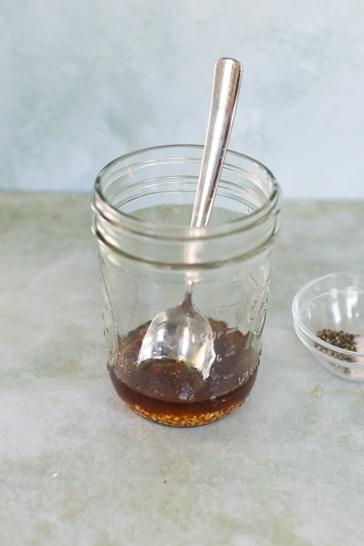 A spoon rests in a glass jar containing brown liquid and seasonings, with a small bowl of pepper set to the side.
