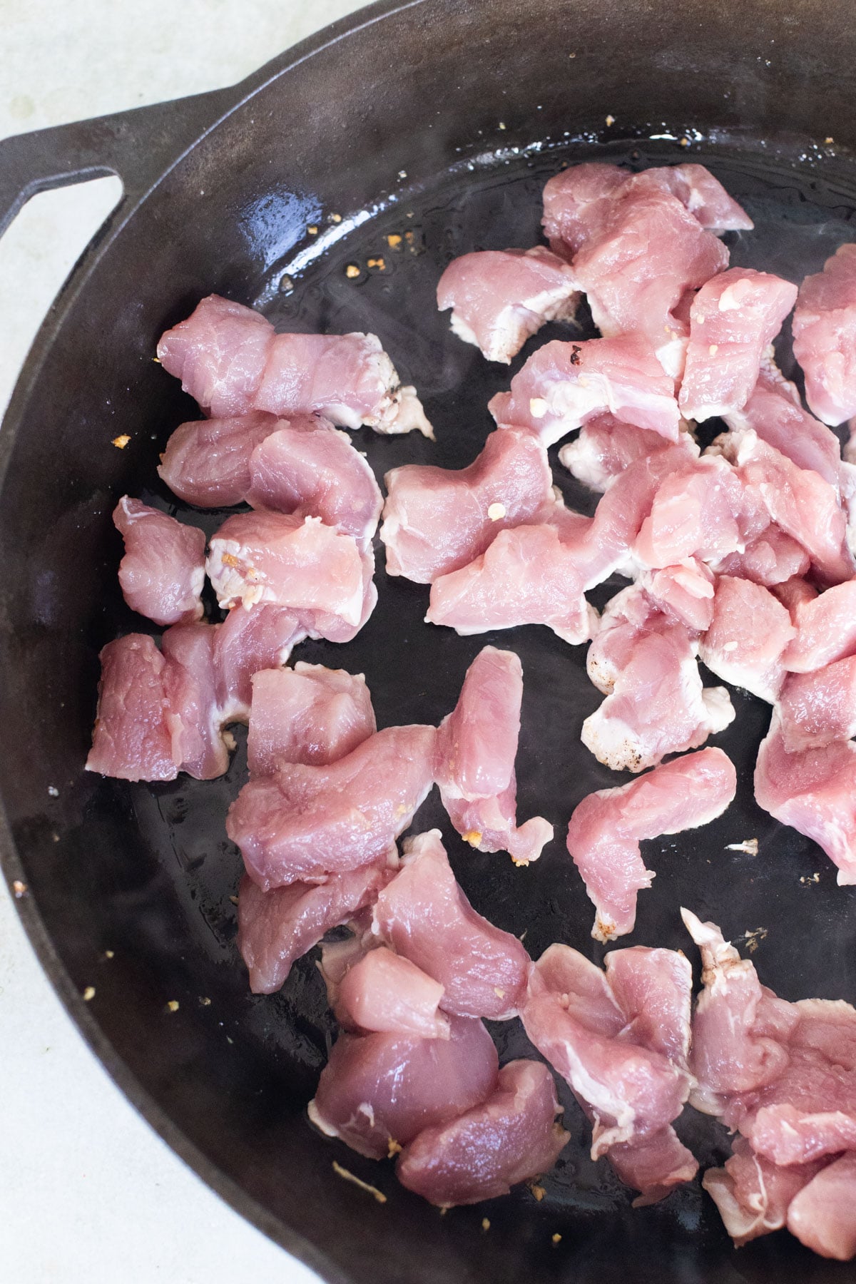 Raw bite sized pieces of pork tenderloin are spread out in a single layer on a black cast iron skillet, ready to be cooked.