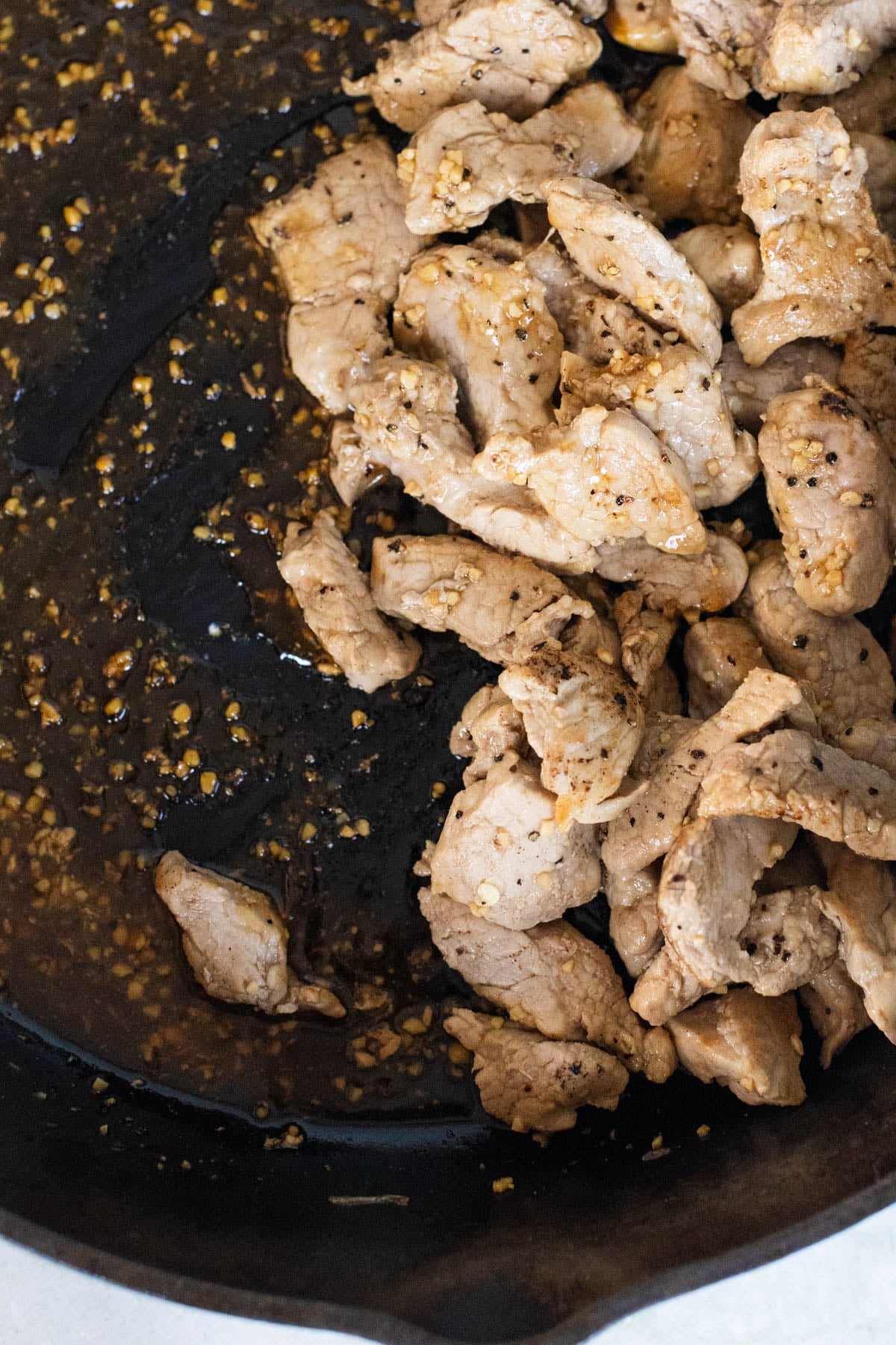 Sliced pieces of cooked pork being stir fried with garlic and seasoning in a cast iron skillet, viewed from overhead.
