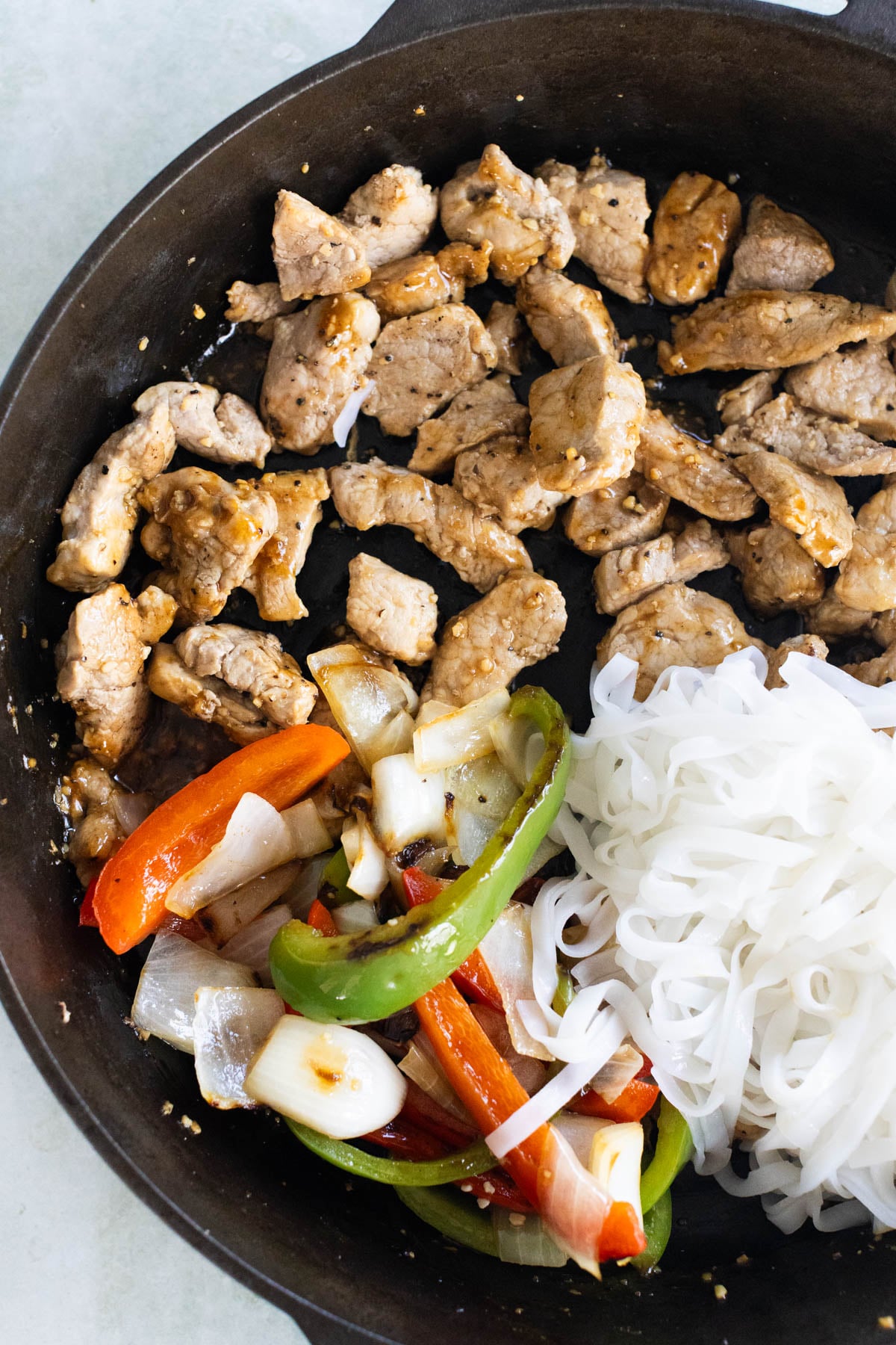A skillet containing cooked sliced meat, stir-fried vegetables including bell peppers and onions, and a portion of cooked rice noodles.