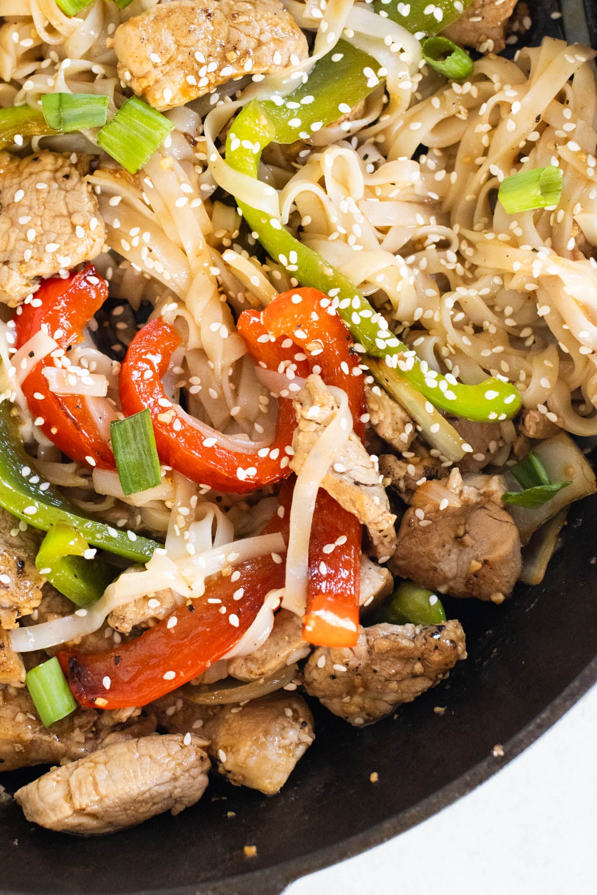 Pork tenderloin stir fry with bell peppers, green onions, and sesame seeds, served in a black skillet over rice noodles.