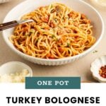 A bowl of turkey bolognese spaghetti with a fork, garnished with herbs, sits on a table next to grated cheese and a glass of white wine.