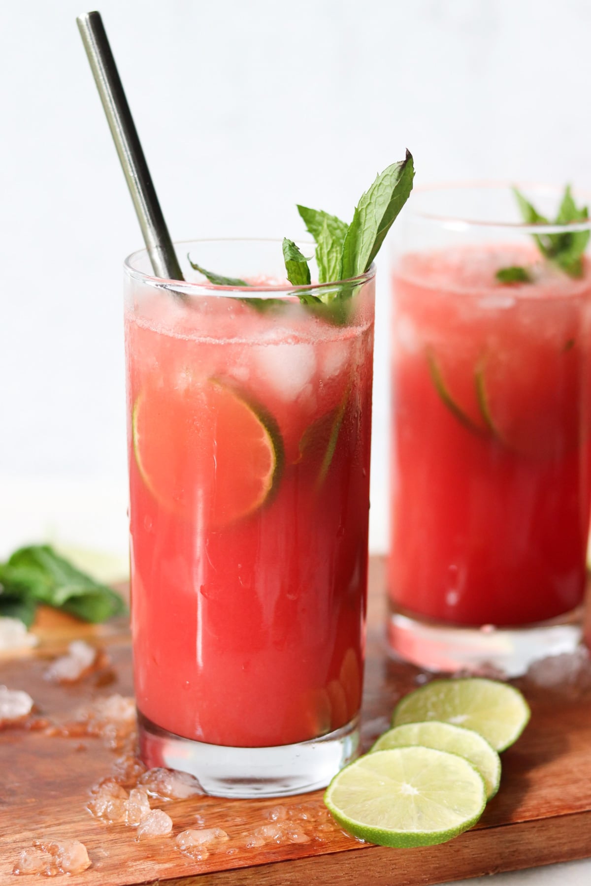 Two glasses of red fruit drink with ice, lime slices, mint sprigs, and metal straws on a wooden board, with lime slices and mint leaves nearby.