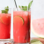 A glass of red beverage with ice, lime slices, and mint garnish, placed on a wooden surface with lime, mint, and ice nearby.