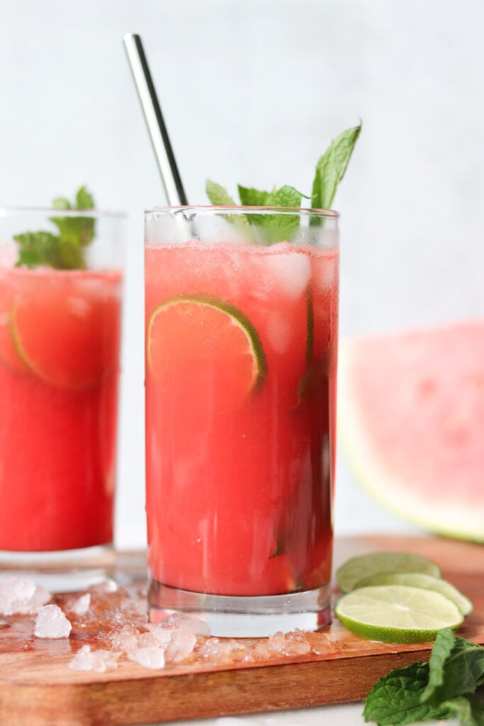 A glass of red beverage with ice, lime slices, and mint garnish, placed on a wooden surface with lime, mint, and ice nearby.