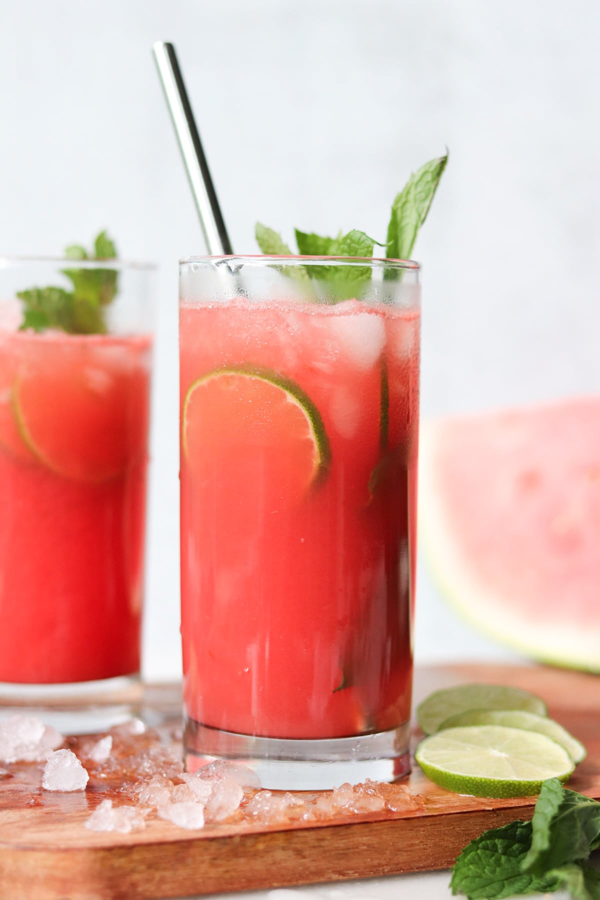 A glass of red beverage with ice, lime slices, and mint garnish, placed on a wooden surface with lime, mint, and ice nearby.