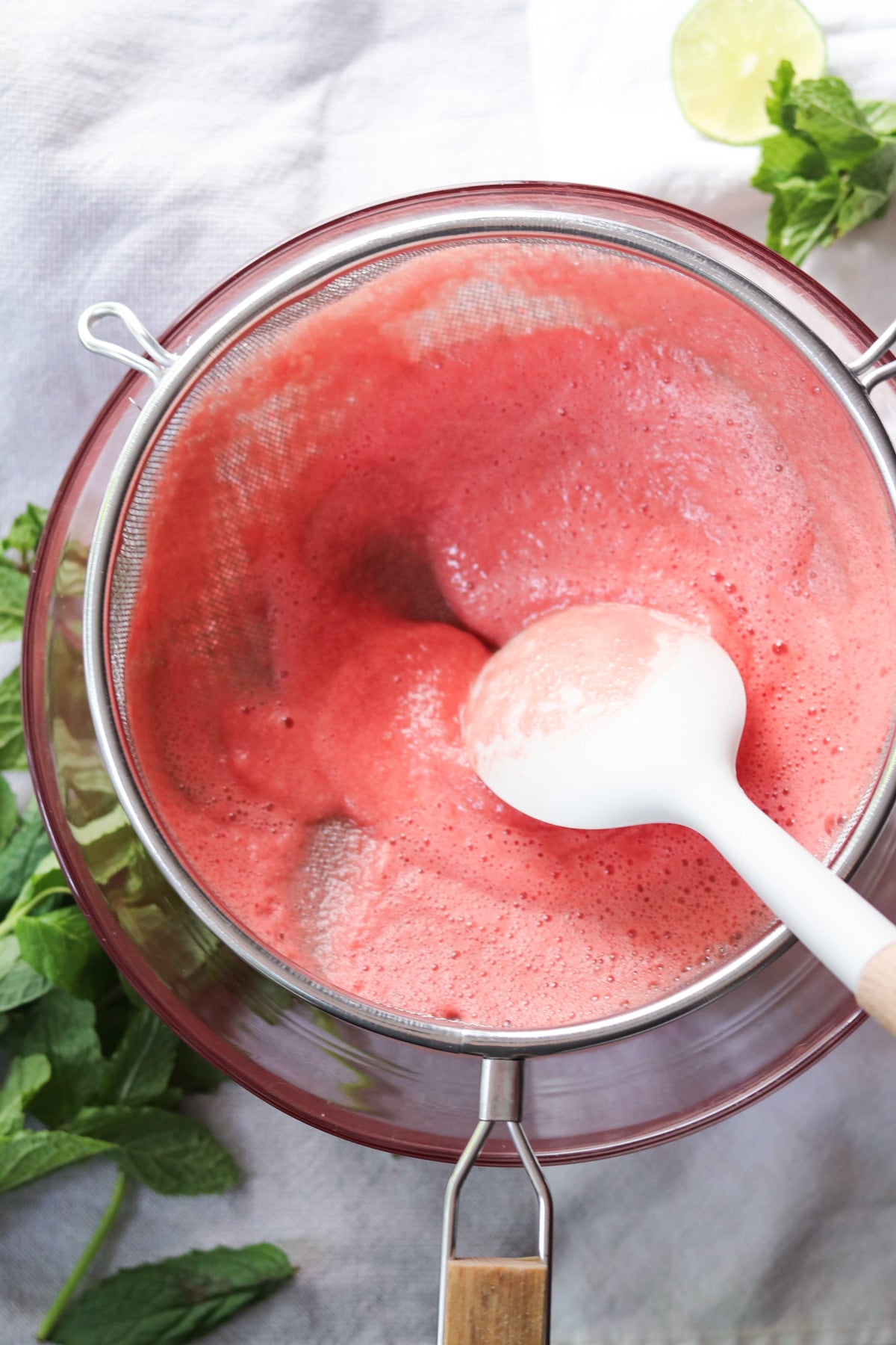 A white spatula presses blended pink fruit puree through a fine mesh strainer over a glass bowl; fresh mint and a lime wedge sit nearby.