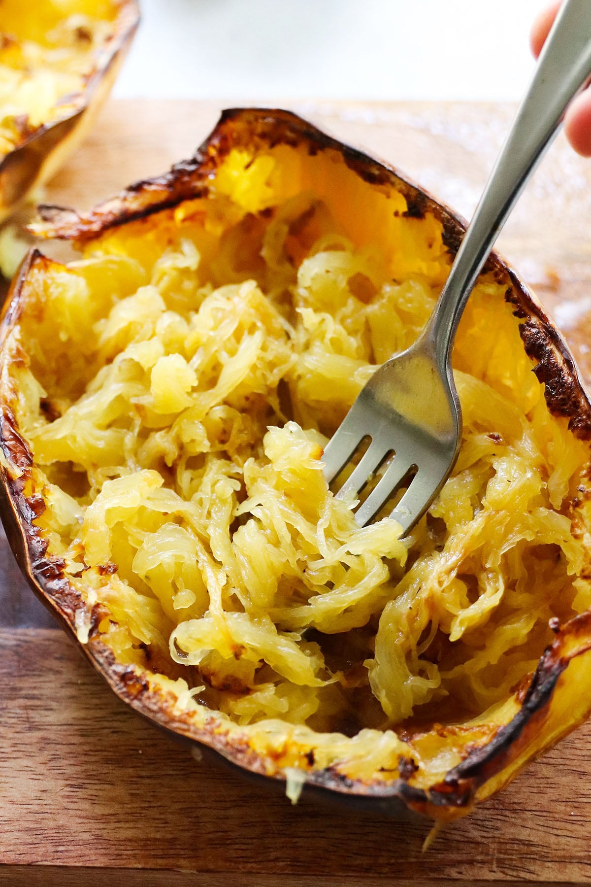 A fork shredding the cooked flesh of a roasted spaghetti squash still in its skin on a wooden surface.