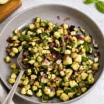 A plate of sautéed diced zucchini and red onion, seasoned with herbs, with a spoon on the side. Nearby are a bottle of olive oil, a bowl of pepper, and fresh basil leaves.
