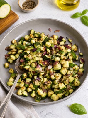 A plate of sautéed diced zucchini and red onion, seasoned with herbs, with a spoon on the side. Nearby are a bottle of olive oil, a bowl of pepper, and fresh basil leaves.