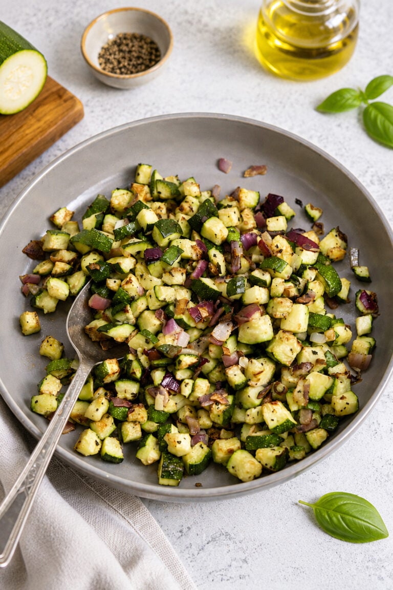 A plate of sautéed diced zucchini and red onion, seasoned with herbs, with a spoon on the side. Nearby are a bottle of olive oil, a bowl of pepper, and fresh basil leaves.