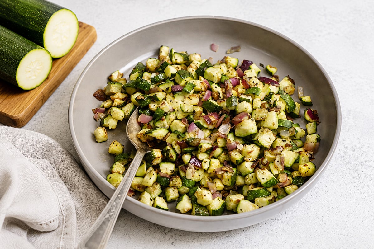 A round plate filled with sautéed diced zucchini and red onion, with a serving spoon resting inside; sliced raw zucchini on a wooden board nearby.