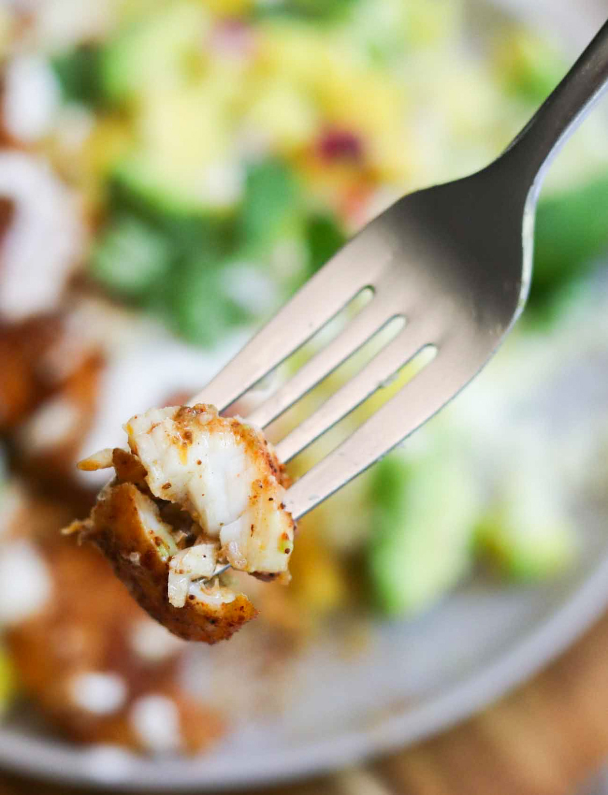 A close-up of a fork holding a bite-sized piece of pan fried flounder, with a blurred plate of food in the background.