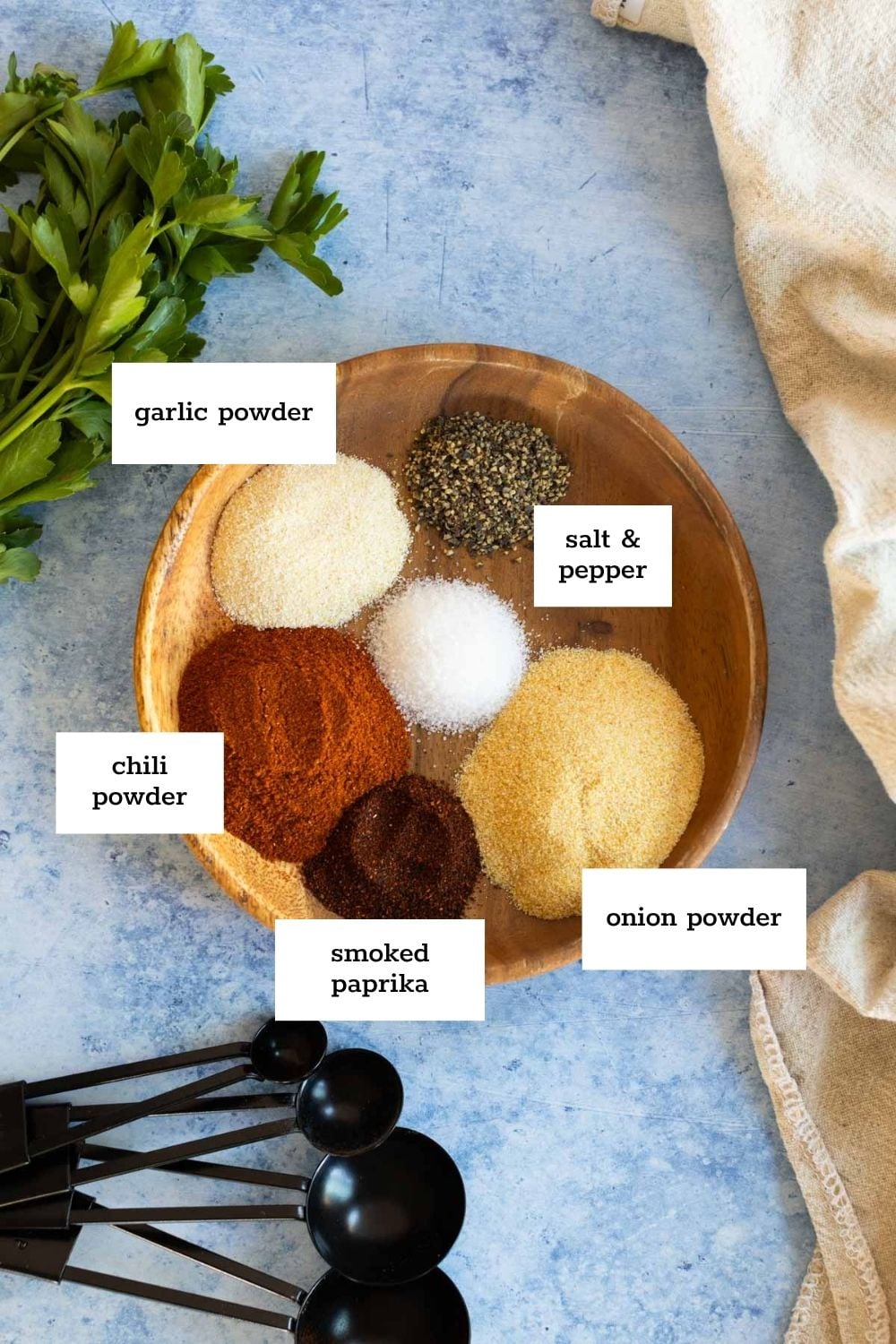 A wooden bowl with garlic powder, chili powder, smoked paprika, onion powder, salt, and pepper, labeled; fresh parsley, measuring spoons, and a cloth are nearby.