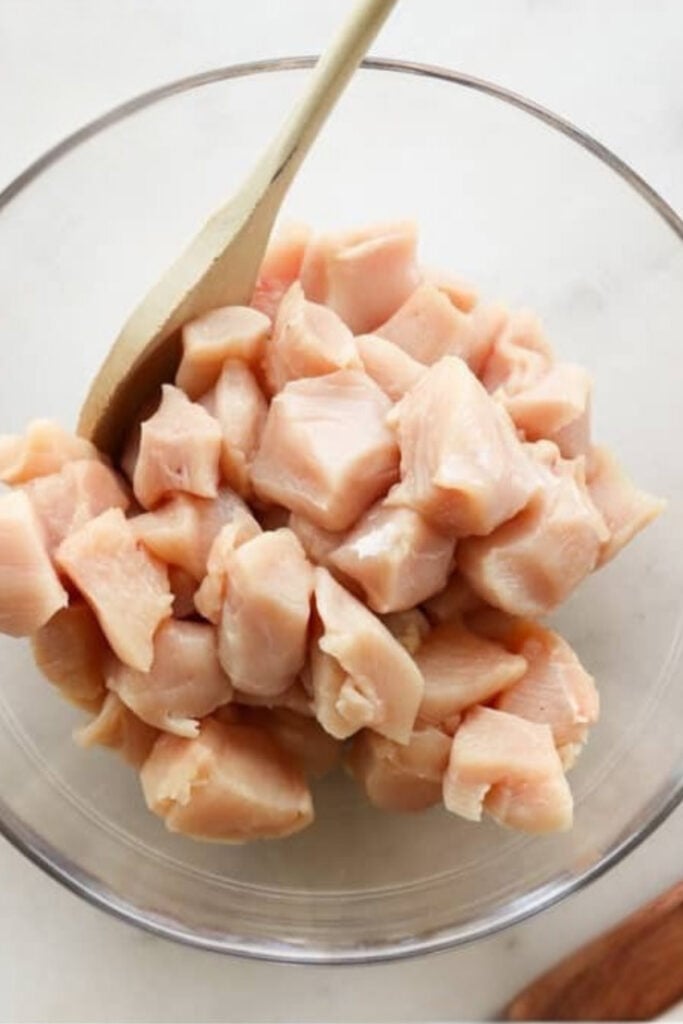 A glass bowl filled with raw, diced chicken pieces being stirred with a wooden spoon.