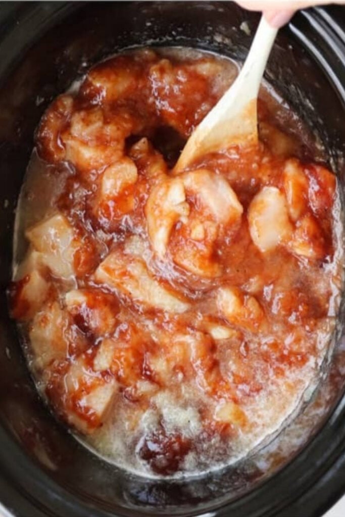 Chunks of raw chicken being stirred with sauce in a slow cooker using a wooden spoon.