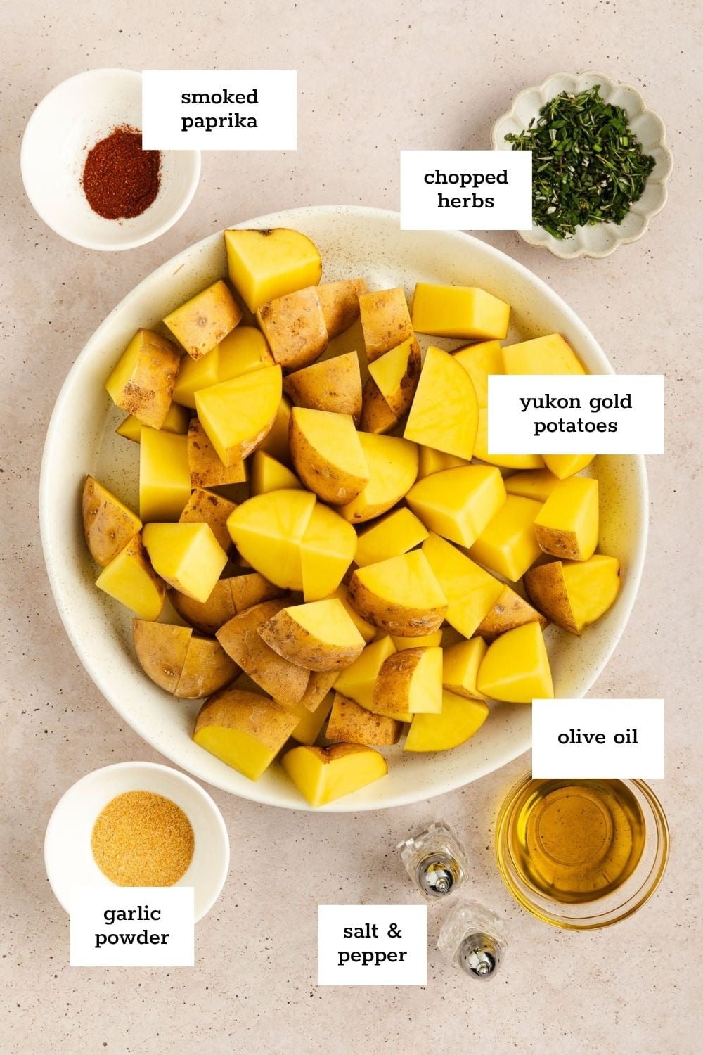 A bowl of chopped Yukon gold potatoes surrounded by small bowls of smoked paprika, chopped herbs, garlic powder, olive oil, and salt & pepper, all labeled.