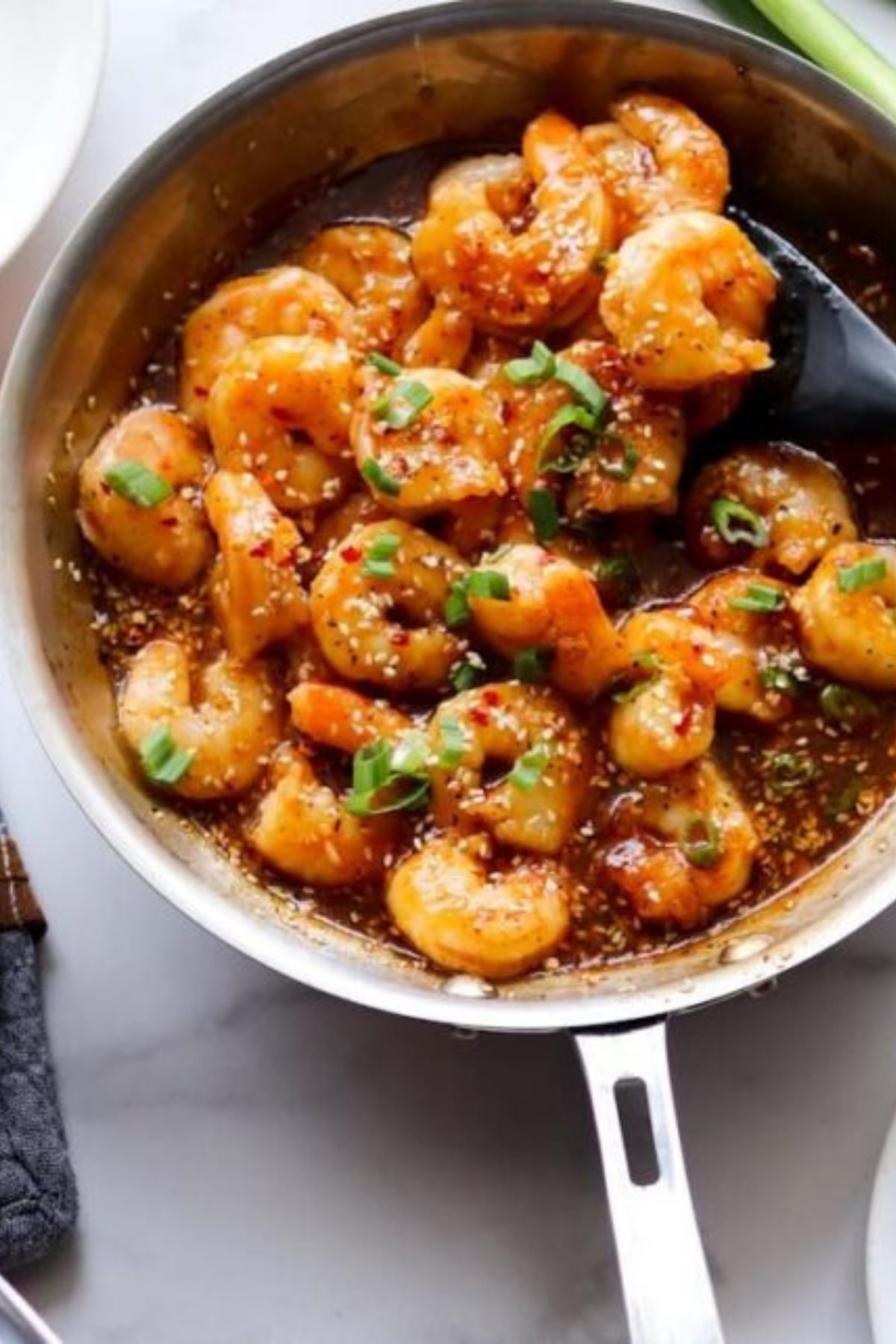 Shrimp cooked in a skillet with a savory sauce, garnished with chopped green onions and sesame seeds, with a serving spoon in the pan.