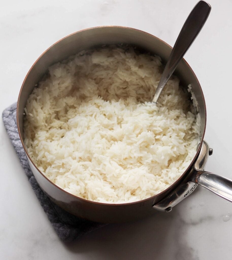 A pot of cooked white rice with a spoon inside, resting on a gray pad on a white surface.