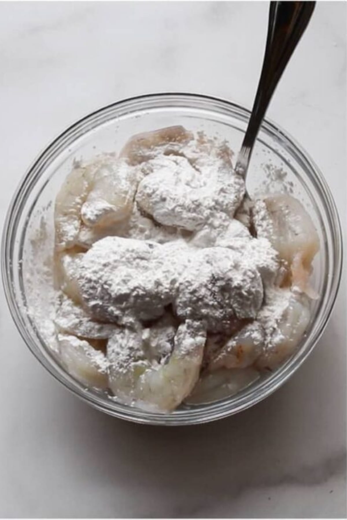 A glass bowl of raw shrimp topped with a white powder, likely cornstarch or flour, with a metal spoon resting inside.
