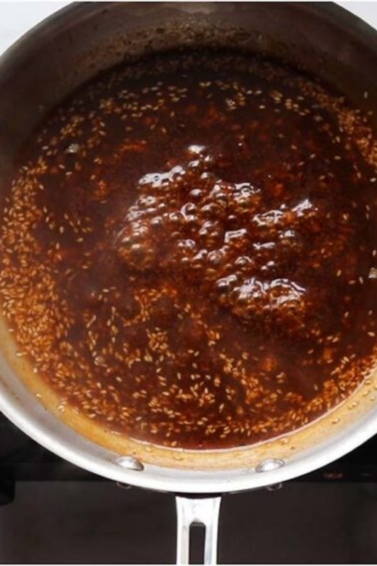 A stainless steel pan on a stovetop contains a dark brown sauce with visible sesame seeds, simmering and bubbling.