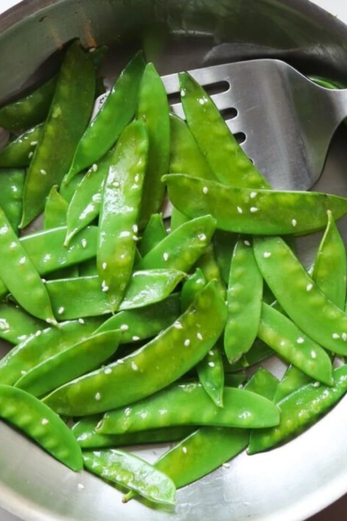 A pan filled with cooked snow peas sprinkled with sesame seeds, next to a metal spatula.