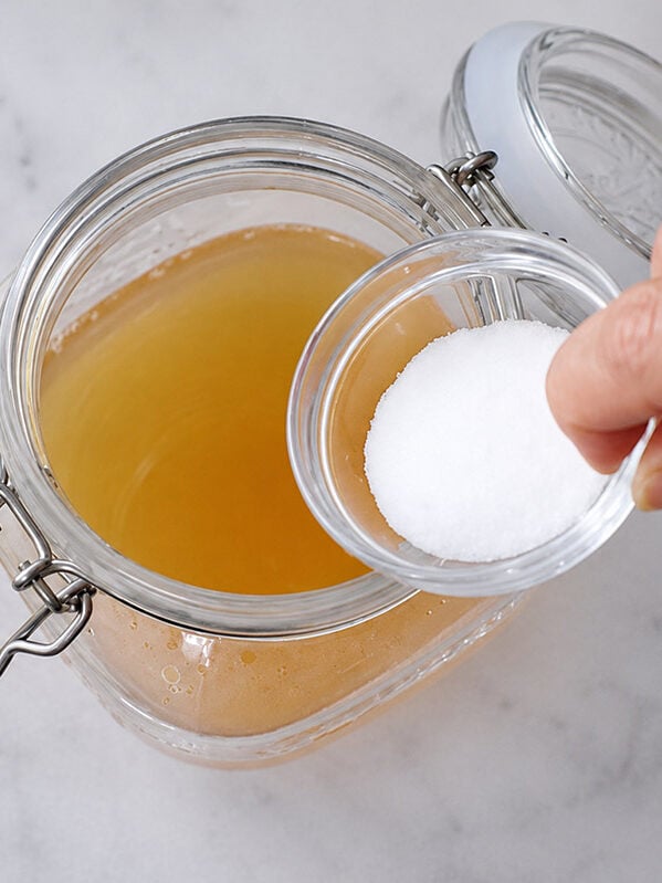 A hand adds a small bowl of white sugar to a glass jar containing a light yellow liquid.