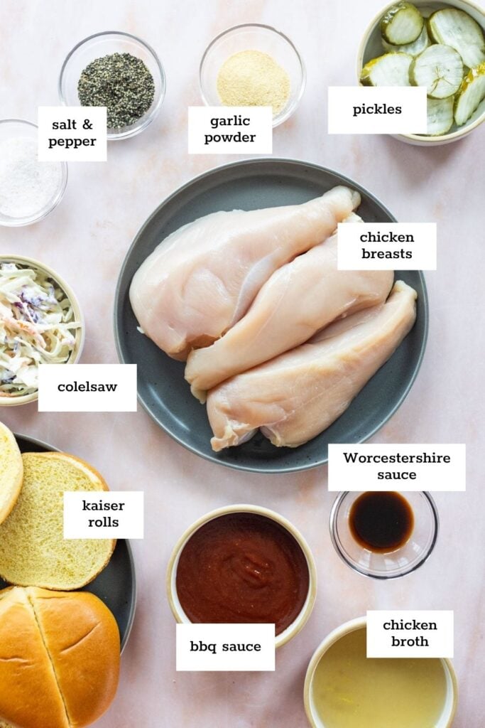 Top-down view of ingredients for a chicken sandwich, including raw chicken breasts, kaiser rolls, coleslaw, pickles, seasonings, Worcestershire sauce, BBQ sauce, and chicken broth.