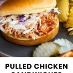 Pulled chicken sandwich topped with coleslaw on a plate, served with potato chips and pickle slices.