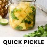 A glass jar filled with pickled pineapple pieces, herbs, and jalapeño slices sits on a counter next to fresh cilantro, a lime, and a pineapple.