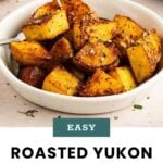 A bowl of roasted Yukon gold potato chunks, seasoned with herbs and pepper, sits on a light-colored surface with a spoon inside.