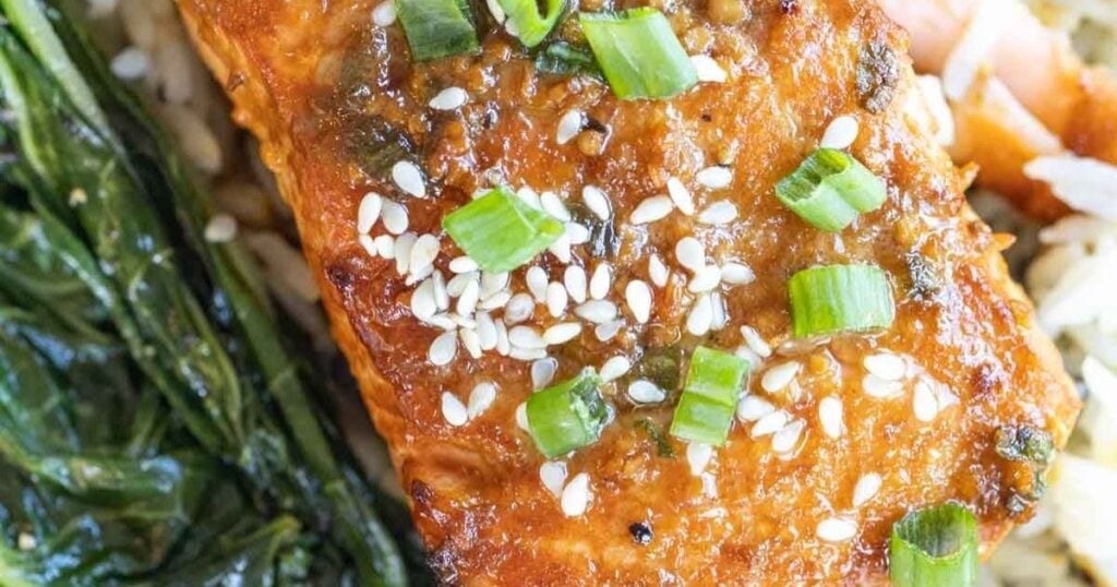 Close-up of a cooked salmon fillet topped with sesame seeds and chopped green onions, served with rice and sautéed greens.