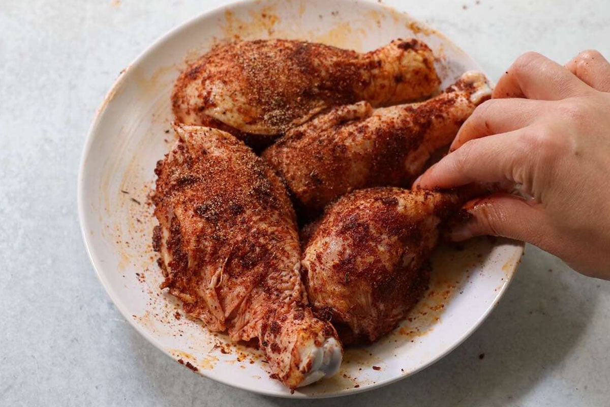 A hand coats raw chicken drumsticks with spices in a white bowl on a light-colored surface, preparing flavorful air fryer chicken legs.