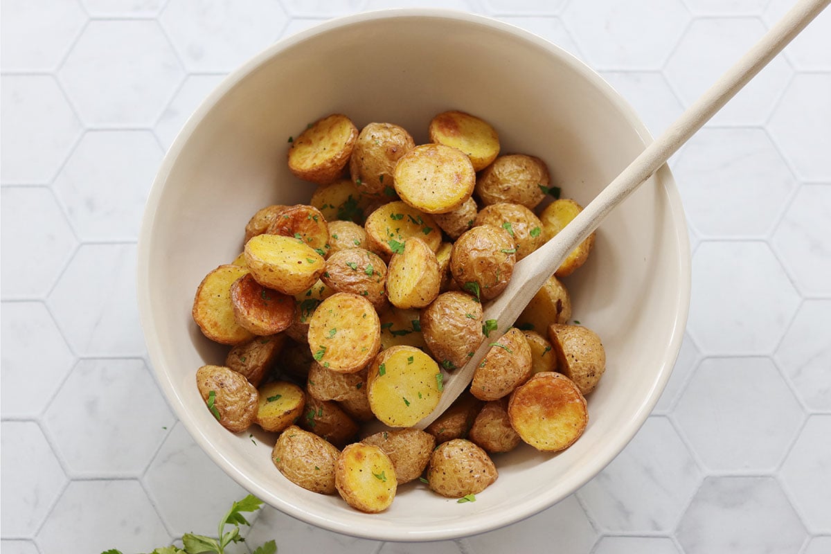 A bowl of roasted baby potatoes with herbs, mixed with a wooden spoon, sits on a white hexagon-tiled surface.
