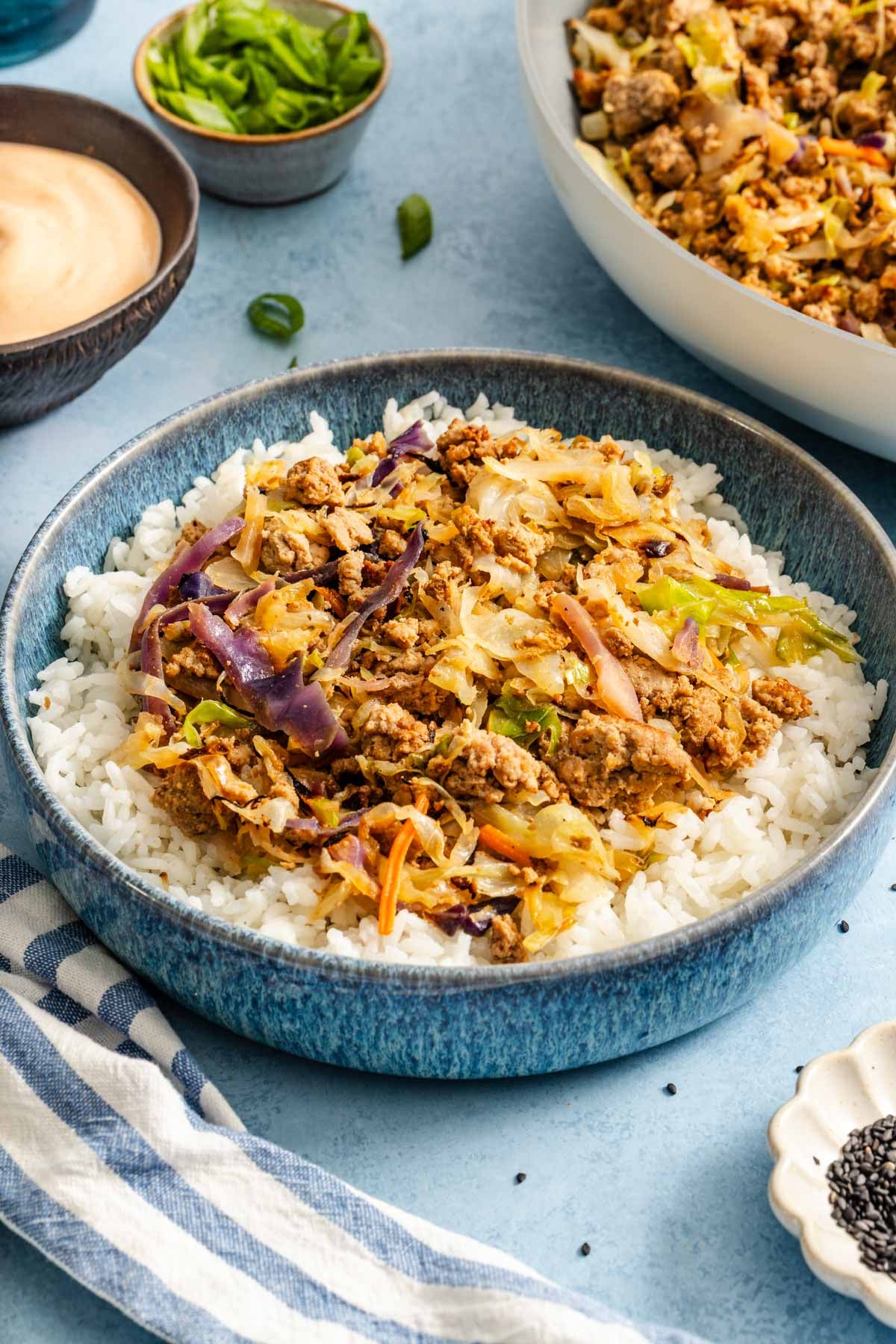 A blue bowl filled with white rice topped with a cooked ground meat and shredded vegetable stir-fry, surrounded by bowls of sauce and sliced green onions.