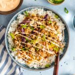 A bowl filled with white rice, ground meat, vegetables, sliced green onions, and a drizzle of creamy sauce, with extra sauce and toppings on the side.