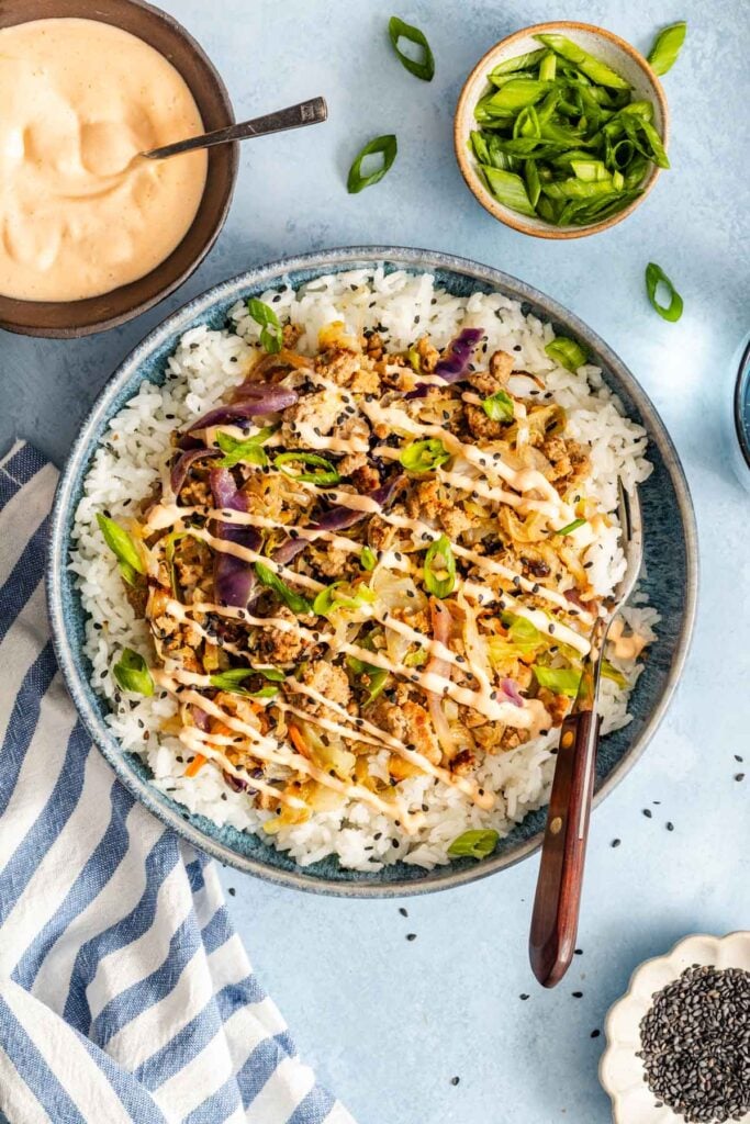A bowl filled with white rice, ground meat, vegetables, sliced green onions, and a drizzle of creamy sauce, with extra sauce and toppings on the side.