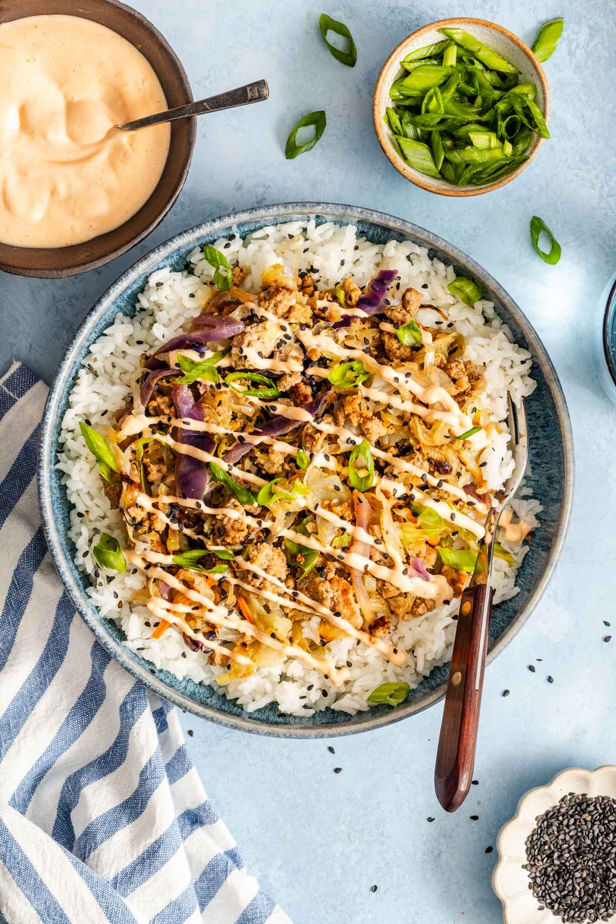 A bowl filled with white rice, ground meat, vegetables, sliced green onions, and a drizzle of creamy sauce, with extra sauce and toppings on the side.