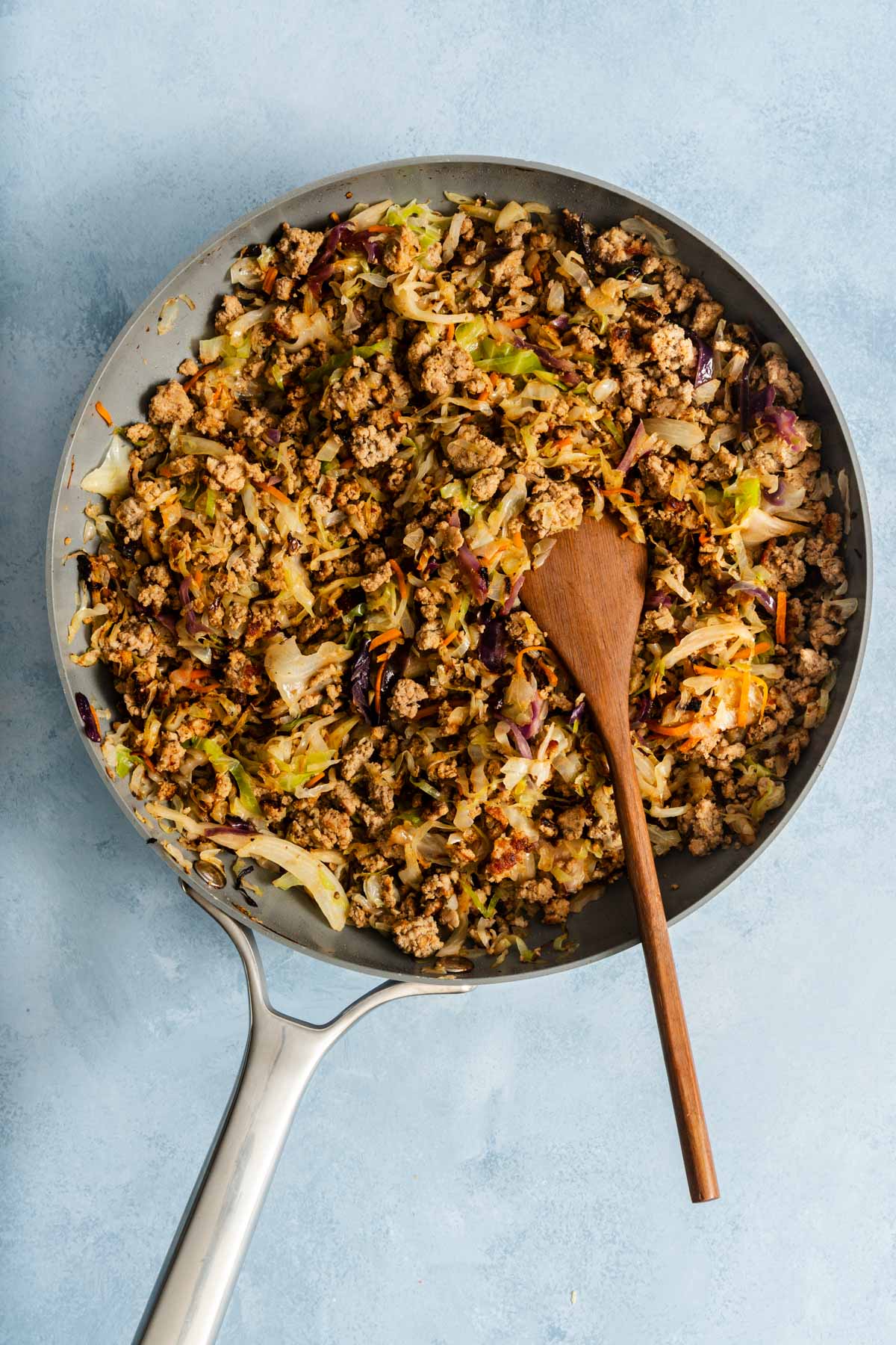 Skillet filled with cooked ground meat, shredded cabbage, and vegetables, with a wooden spoon resting inside. The background is a light blue surface.