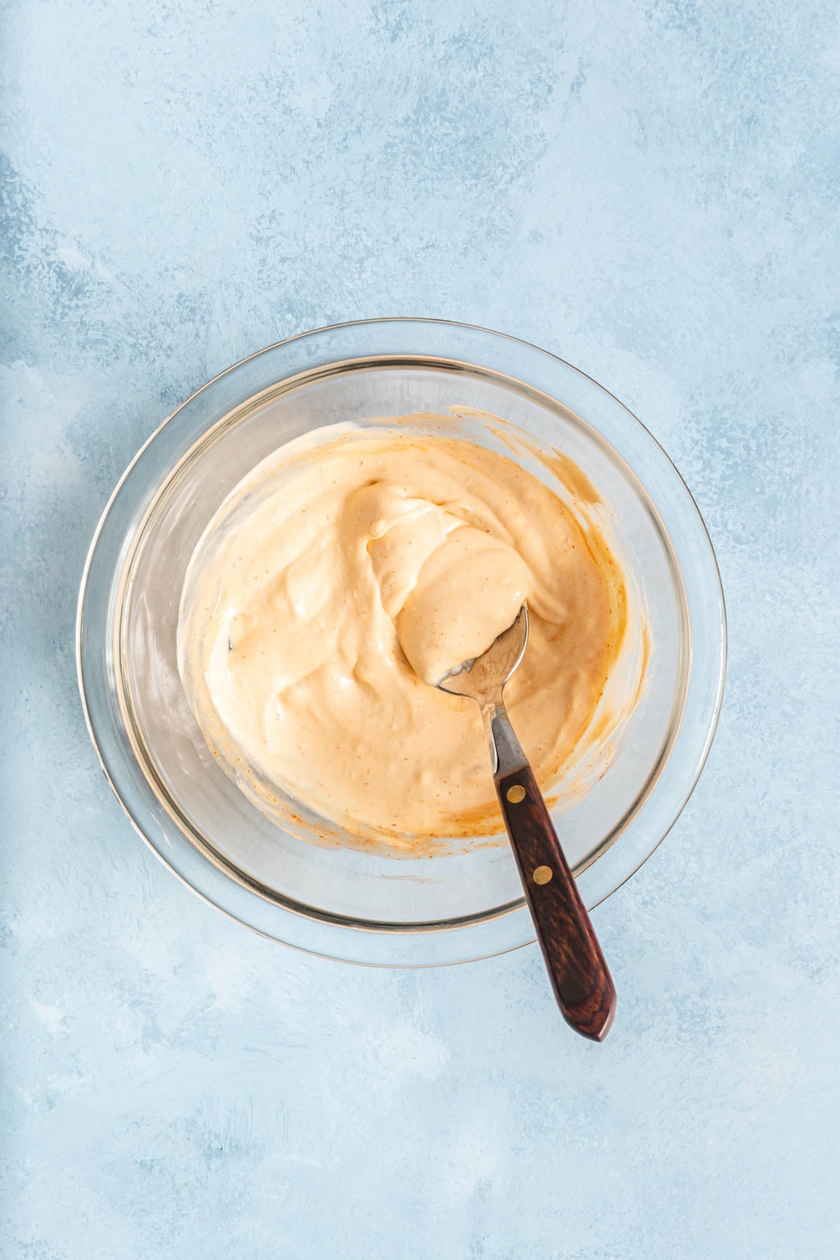 A glass bowl of creamy, light orange sauce with a spoon inside, placed on a light blue textured surface.