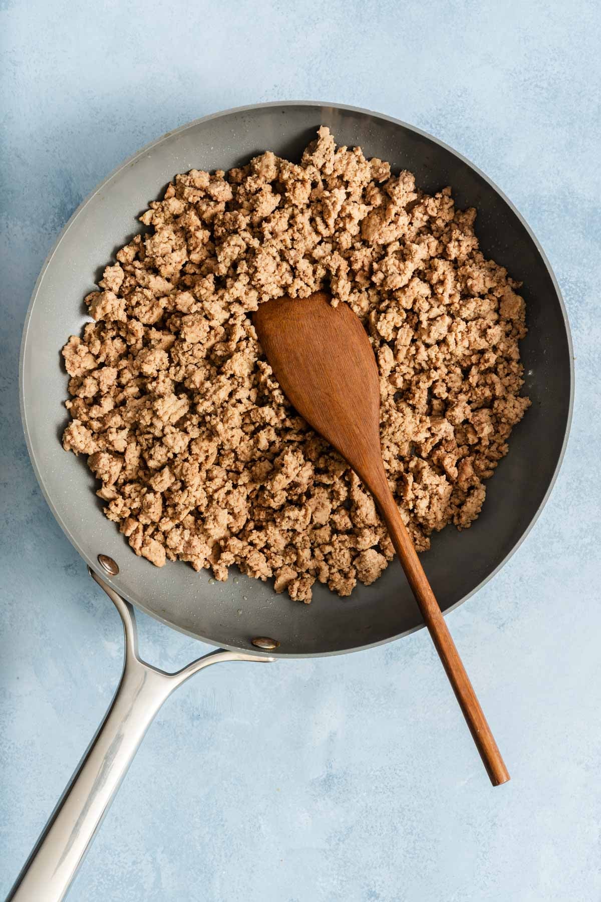 Cooked ground turkey in a skillet with a wooden spoon on a light blue surface.