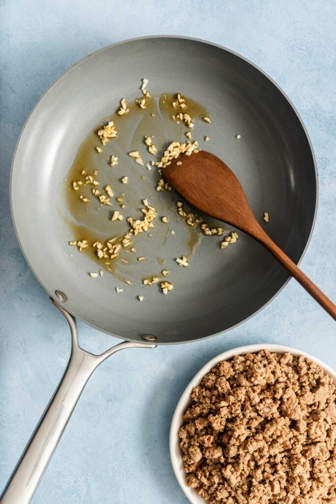 A frying pan with sautéed garlic and a wooden spoon sits next to a bowl of cooked ground meat on a light blue surface.