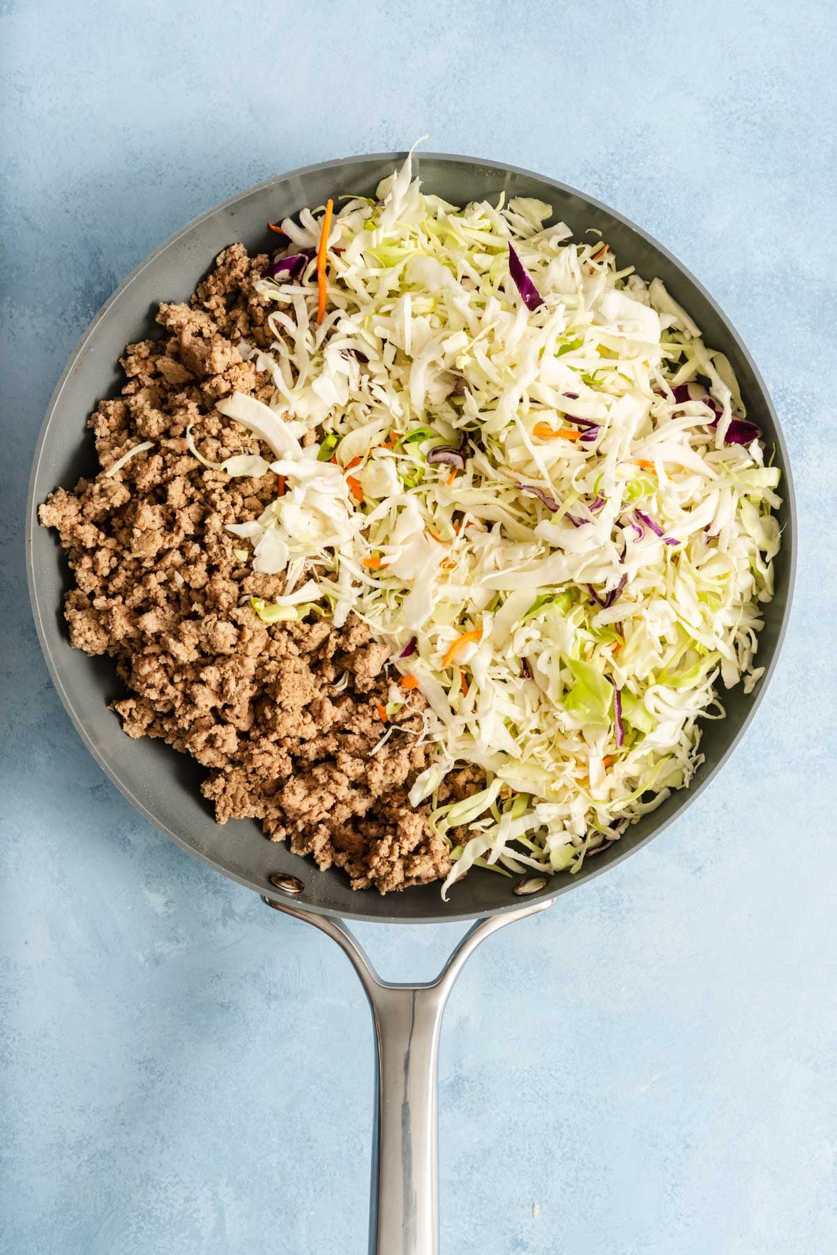 A skillet containing cooked ground turkey on one side and shredded cabbage and carrots on the other, for an egg roll in a bowl dinner.