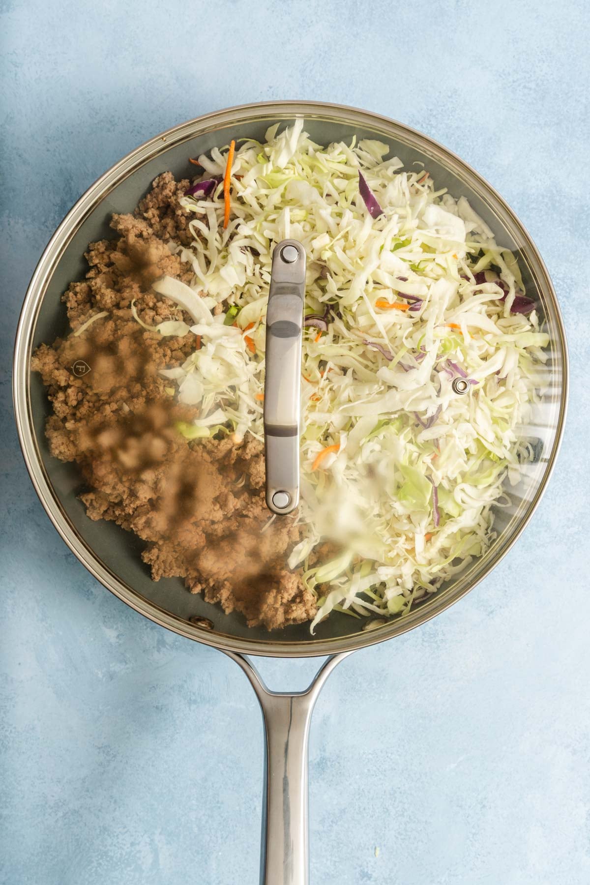 A frying pan with ground meat and shredded cabbage mixture cooking under a glass lid on a light blue surface.
