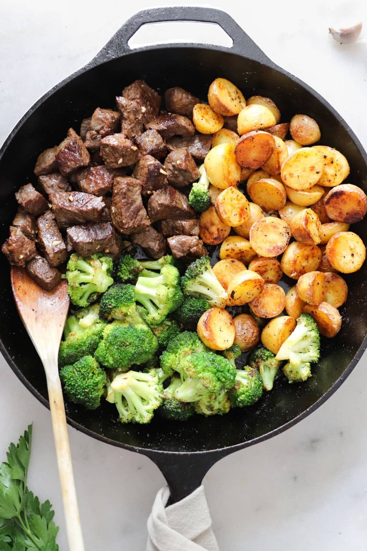A cast iron skillet with cooked steak cubes, seasoned roasted potatoes, and steamed broccoli, arranged in sections with a wooden spoon on the side.