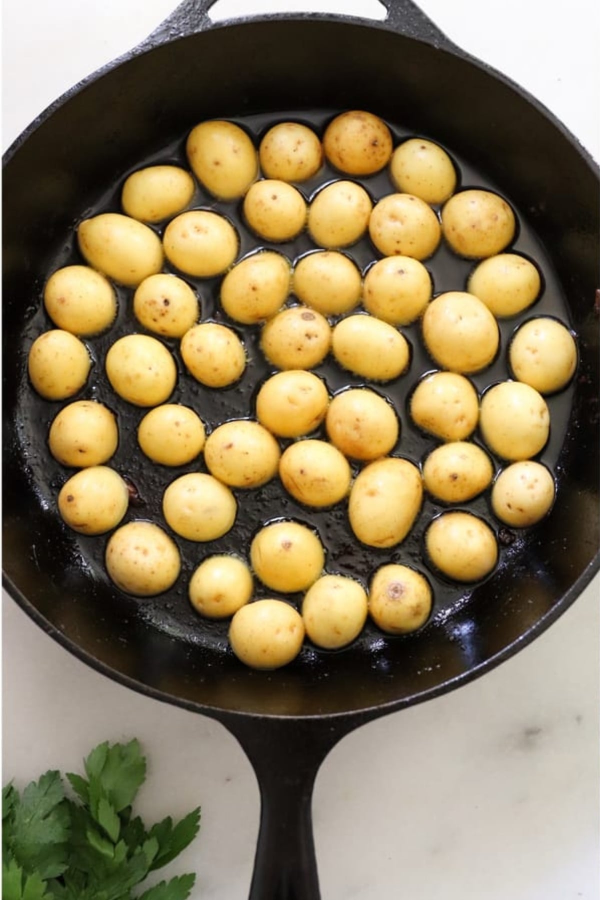 Small yellow potatoes arranged in a single layer in a black cast iron skillet with parsley leaves nearby on a white surface.