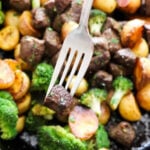 A fork holds a piece of cooked beef above a skillet filled with beef, roasted potatoes, and broccoli.