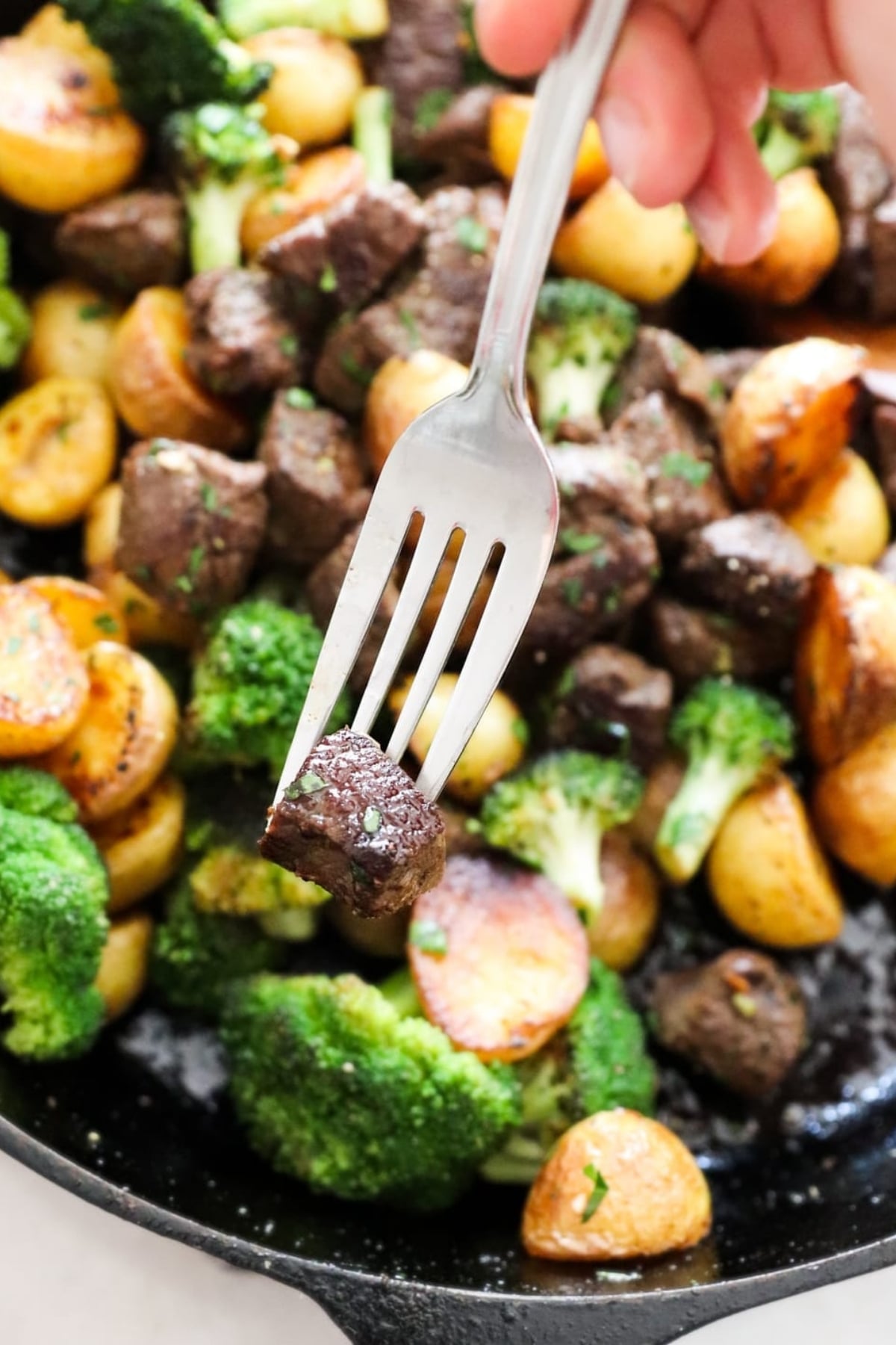 A fork holds a piece of cooked beef above a skillet filled with beef, roasted potatoes, and broccoli.