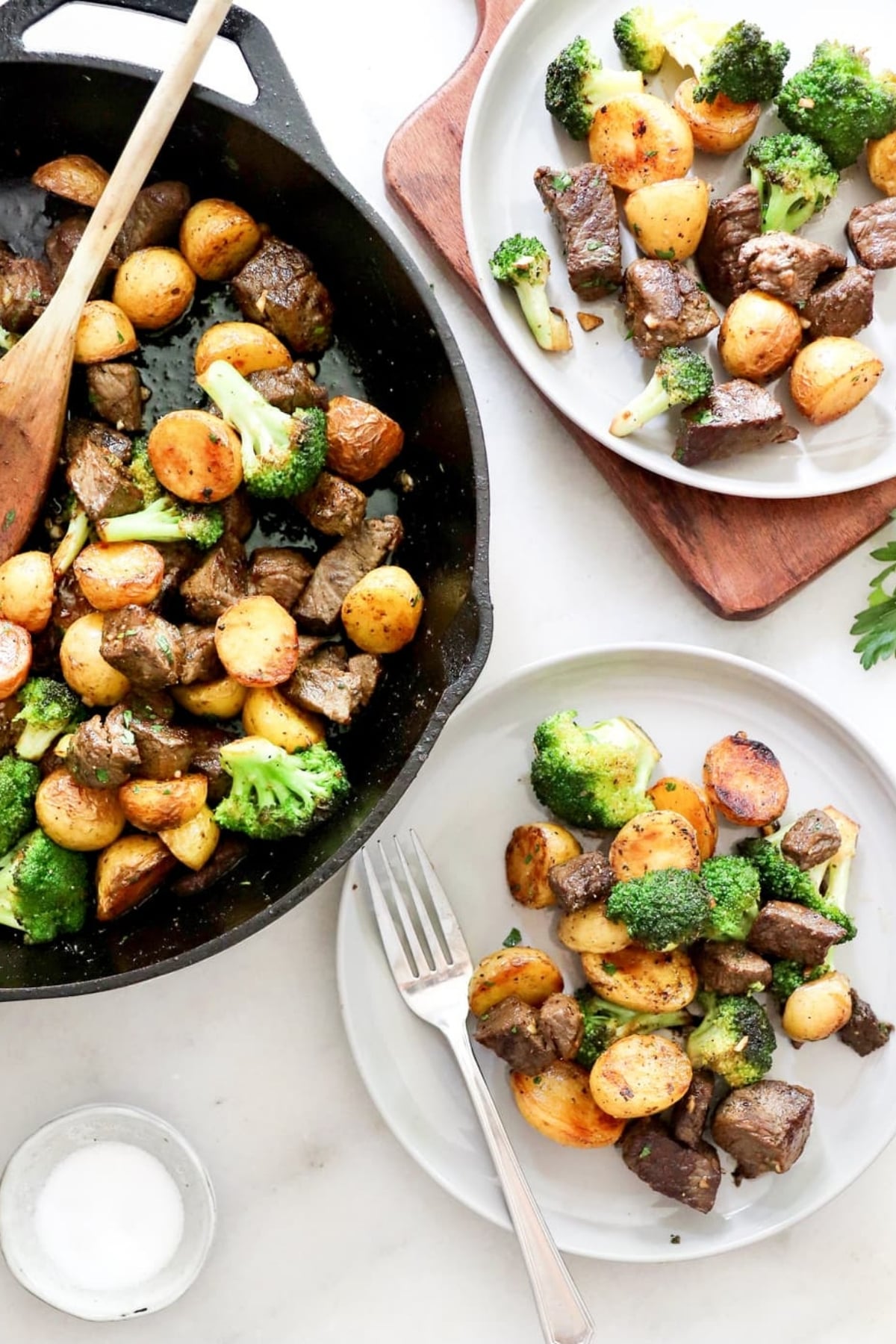 A skillet and two plates with cooked beef, baby potatoes, and broccoli, with a wooden spoon in the skillet and a fork on one plate. A small bowl of salt is nearby.